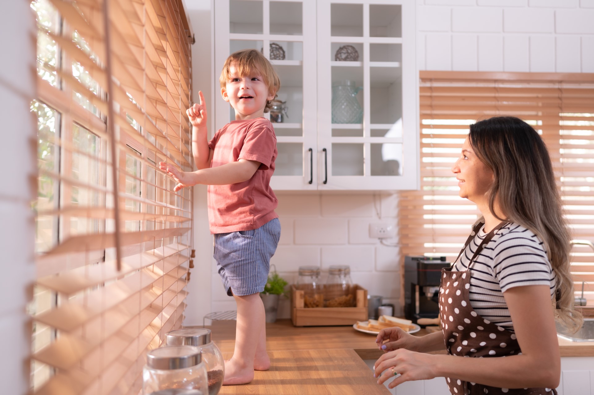 A mother playing with her child on the kitchen counter as he looks through the blinds.