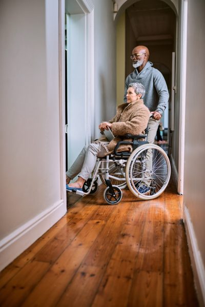 A man pushing a senior woman around her home in a wheelchair.